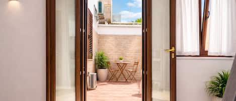 Habitación con puerta corrediza de vidrio que da a un patio exterior con mesa y sillas, una planta en maceta y un muro de piedra.