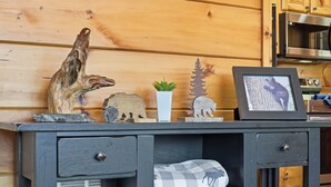 Interior de una cabaña de madera con un escritorio que muestra un cuadro enmarco, una pequeña planta en maceta y varios objetos decorativos.