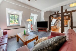 Un salon spacieux avec une télévision, une table basse en bois, un canapé rouge et une vue sur la cuisine grâce à une porte ouverte.