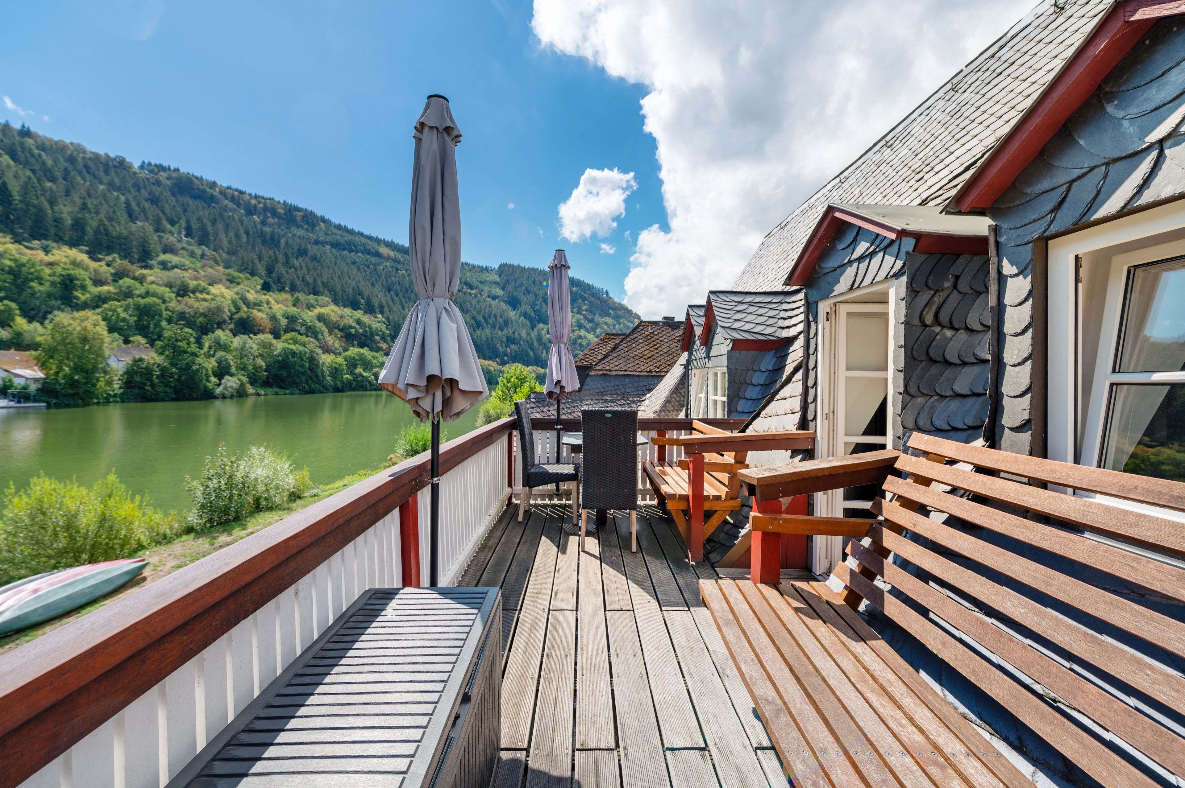 Eine Holzterrasse mit Sitzgelegenheiten und Sonnenschirmen mit Blick auf einen See und bewaldete Hügel.
