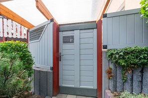 Une porte grise avec une poignée en métal, une petite enseigne au-dessus, et une clôture blanche en bois avec des plantes vertes de chaque côté.