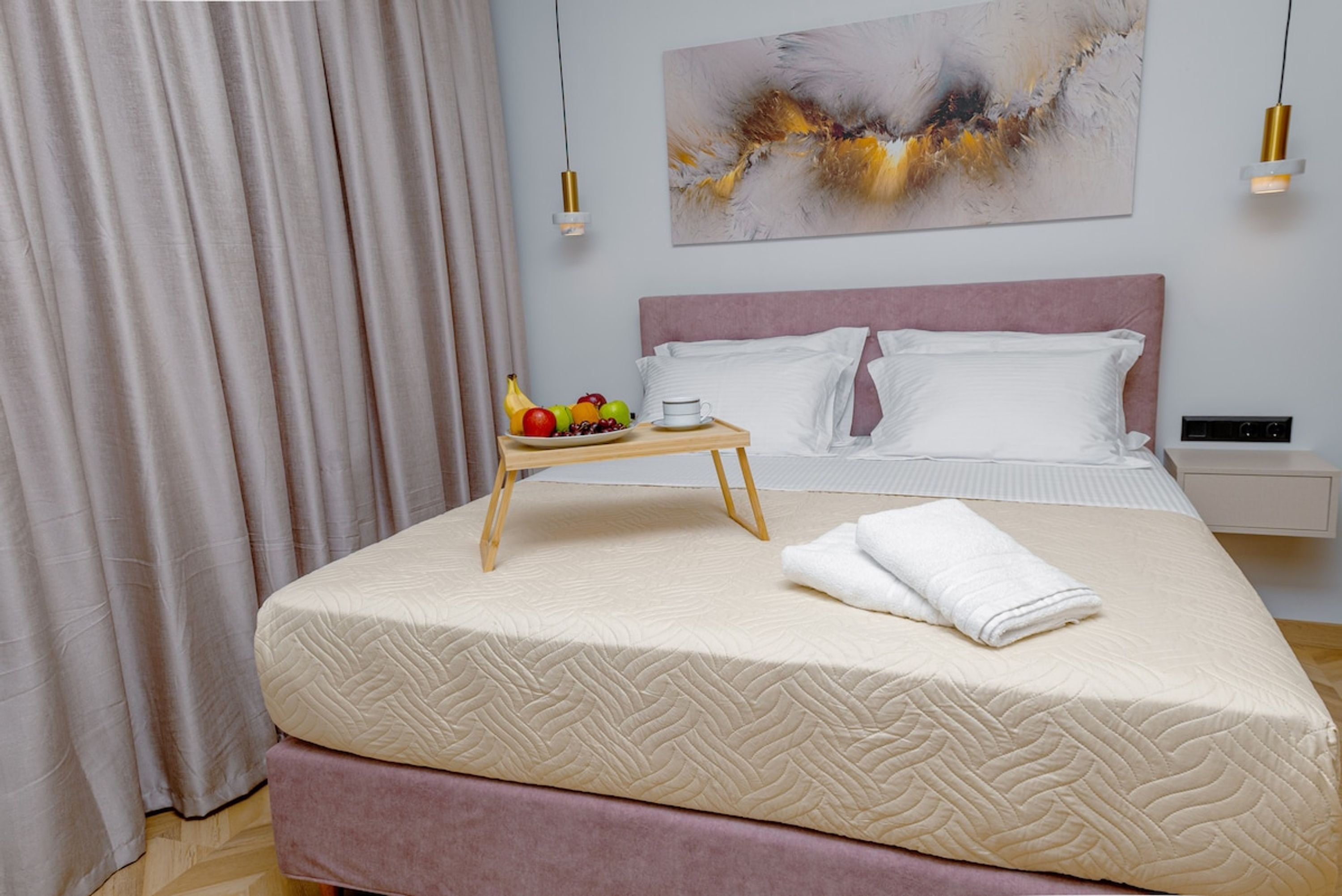A neatly made bed with a pink headboard, a bedside table with a tray of fruit and a cup, and a modern abstract painting on the wall.