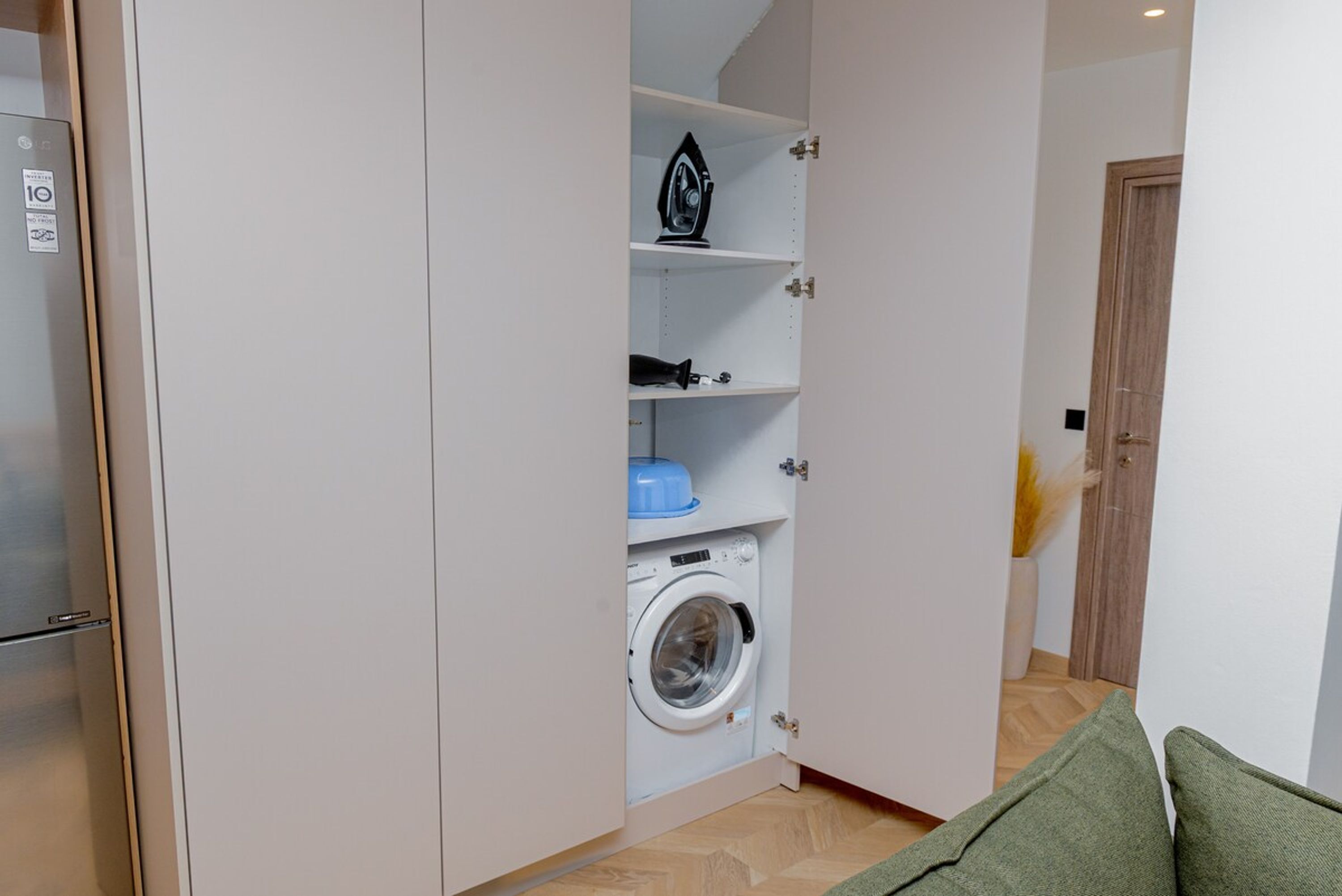 Pieni ja käytännöllinen pyykinhuoltotila, jossa on pyykinpesukone, hyllyjä, joilla on vedenkeitin ja sininen säilytysastia, sekä taustalla puinen ovi.