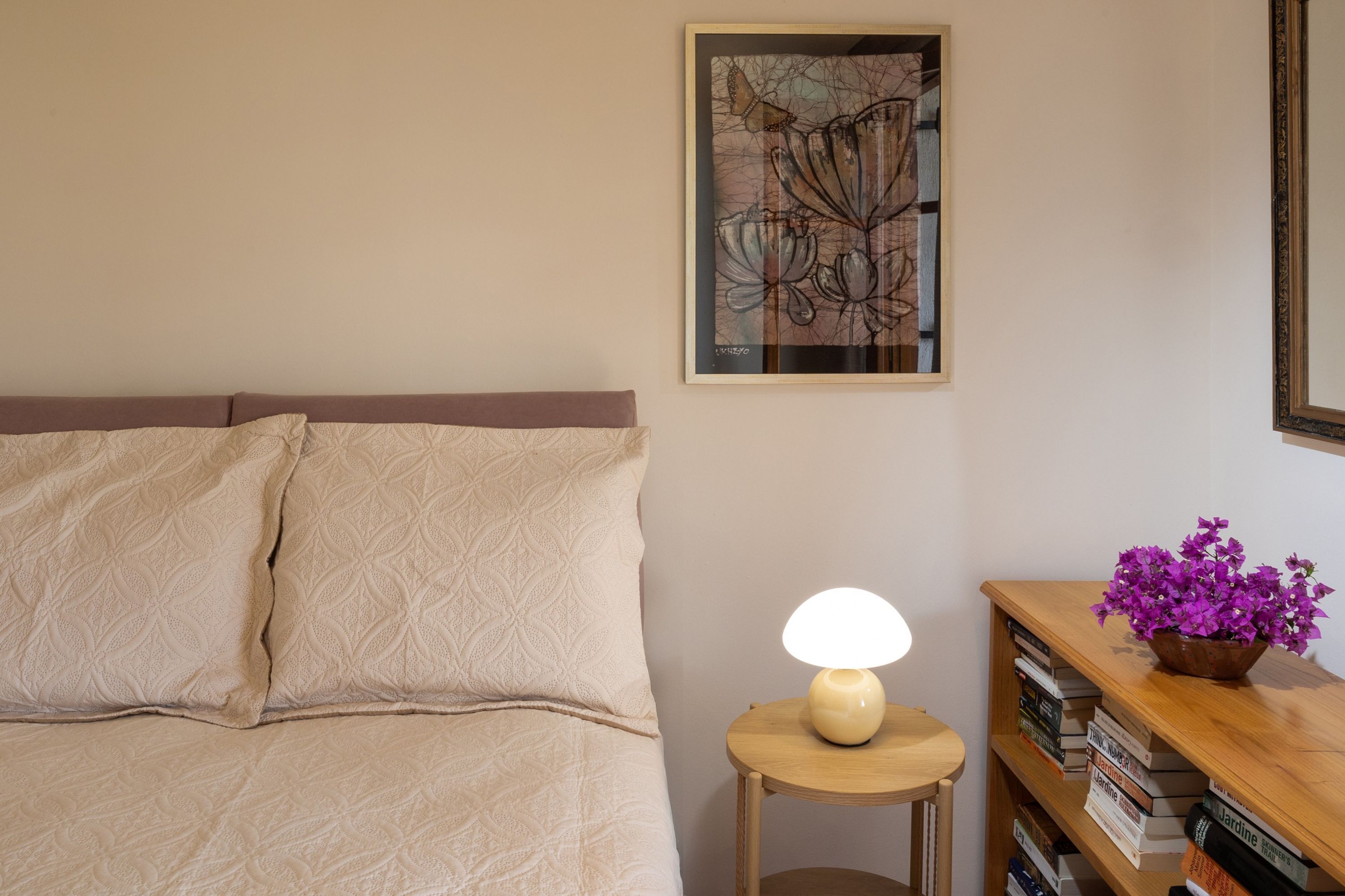 Une chambre à coucher avec un lit, une table de chevet avec une lampe, une œuvre d’art encadrée au mur et une bibliothèque en bois remplie de livres.