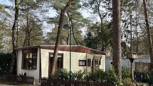 Una cabaña pequeña de un piso con fachada blanca y detalles en marrón, rodeada de árboles y una valla de madera.