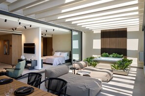 A modern, well-lit living area with a sofa, dining table, and a large bed. There is a small table with two glasses and a candle. The room has a large window and a view of the outdoors.