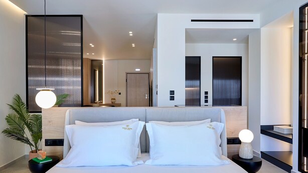 A modern hotel room with a large bed, a bedside table with a lamp, a potted plant, and a mirror reflecting the room. - Executive Apartment by Fasma Suites (Pireas)
