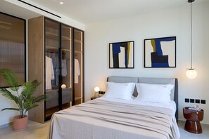 A modern hotel room with a large bed, a wardrobe, two abstract paintings, a potted plant, and a bedside table with a lamp.