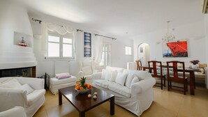 A living room with white furniture, a fireplace, a dining area with wooden chairs, and a large window with curtains. - Naxos Kastraki Homey Villa with Sea View (Kastraki)