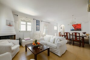 A living room with white furniture, a fireplace, a dining area with wooden chairs, and a large window with curtains. - Naxos Kastraki Homey Villa with Sea View (Kastraki)