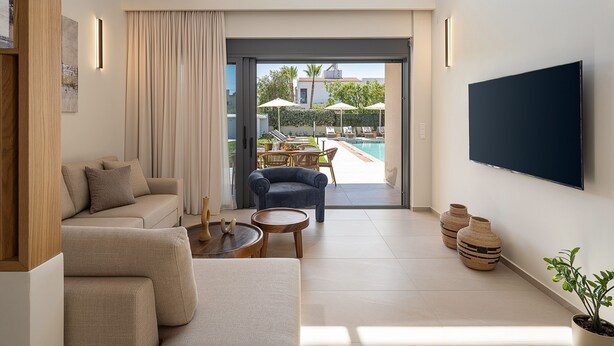 Un soggiorno moderno con divano, poltrona e tavolino, una TV a parete e vista su una zona piscina esterna.
