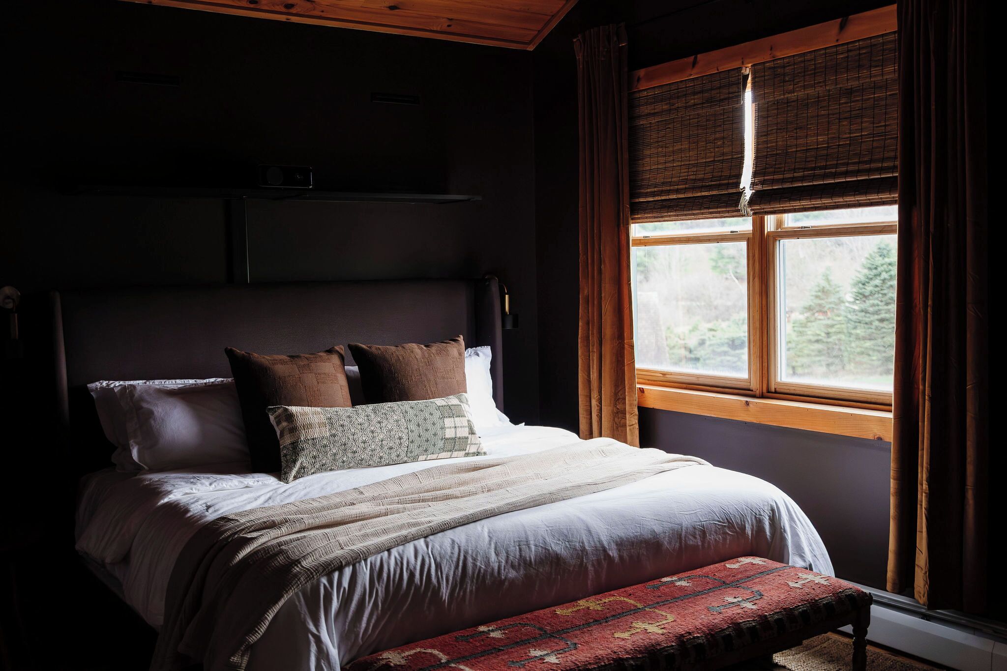 Une chambre à coucher avec un lit, des oreillers, un banc, une fenêtre avec des rideaux et une tête de lit.