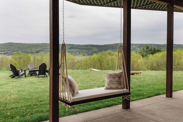 Un fauteuil suspendu avec des coussins, une table de pique-nique et des chaises, le tout installé dans un espace extérieur verdoyant avec vue sur les collines.