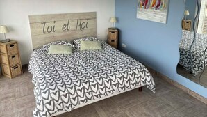 Un dormitorio con cama, dos mesitas de noche, un espejo adosado a la pared, un cuadro y un cabecero de madera con texto.
