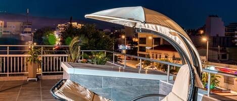 Une terrasse sur le toit, dotée d’un espace salon avec des coussins, d’un auvent et offrant une vue sur le paysage urbain la nuit.
