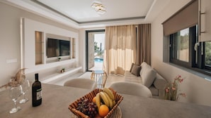A modern living room with a sofa, a dining table with wine glasses and a fruit basket, a TV mounted on the wall, and a view of an outdoor pool. - J&K Villa with private pool (Zakynthos)