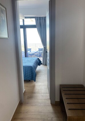 Hotellihuone, jossa on puulattia, sinisellä peitteellä varustettu sänky, penkki ja näkymä ulkoiselle parvekkeelle.