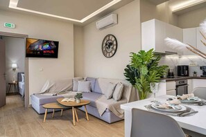 A modern living room with a grey sofa, a coffee table, a TV displaying Netflix, a wall clock, and a dining area with a table set for two. - Pietra Cave Suite with private pool (Zakynthos)