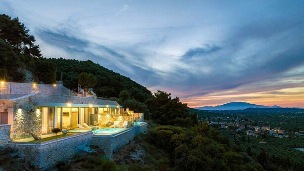 A stone villa with a swimming pool, overlooking a mountainous landscape at dusk.