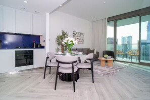 A modern living room with a dining area, a kitchen with white cabinets and a blue countertop, and large windows offering a city view. - Luxury 1BR Burj+Canal View - Heart of Business Bay (Dubai)