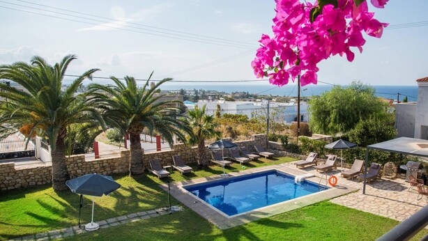 A swimming pool with lounge chairs and umbrellas, surrounded by palm trees and a view of the sea. - Villa Rafaella in Rethymno (Rethymno)