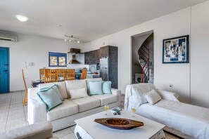 A modern living room with a sofa, a coffee table, and a bed. There is a kitchen area with cabinets and a stove. A blue door and a staircase are visible. - Elounda Senses Luxury villa with private pool (Agios Nikolaos Municipality)