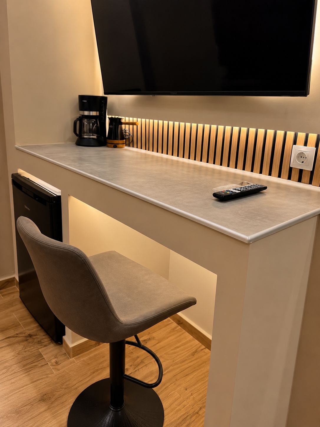A modern kitchenette with a built-in coffee maker, a remote control on the countertop, and a TV mounted above.