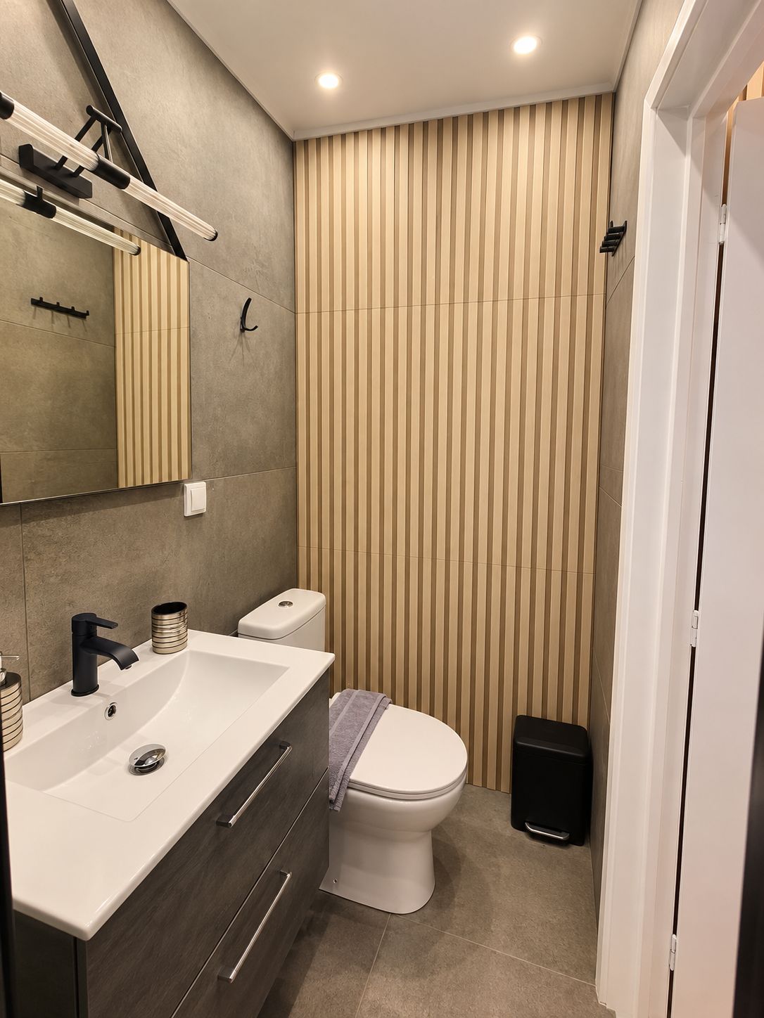 Ein modernes Badezimmer mit einem großen Spiegel, einem weißen Waschbecken, einer Toilette und einer Wand mit vertikalen Streifen.