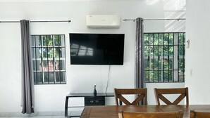 A modern living room with a flat-screen TV mounted on the wall, a wooden dining table with chairs, and two windows with curtains. - 2-bedrooms next to Grace Bay (Grace Bay)