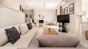 Ein modernes Wohnzimmer mit einer Couch, Couchtisch und Fernseher. An den Wänden hängen Bilder in Rahmen, und im Hintergrund ist ein Kamin zu sehen.