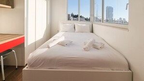Ein Hotelzimmer mit einem Bett, zwei Kissen und zwei gerollten Handtüchern. Ein Fenster bietet Blick auf die Stadt.