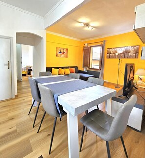Uma sala de estar moderna com uma mesa de jantar, cadeiras cinzas, um sofá com almofadas amarelas, uma TV e obras de arte nas paredes.