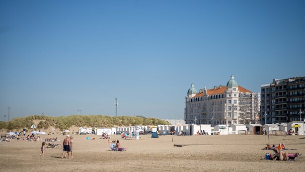 Ein Strand mit Zelten und Sonnenschirmen, ein großes Gebäude mit einer Kuppel und ein Sandbereich mit Menschen.