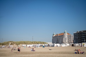 Ein Strand mit Zelten und Sonnenschirmen, ein großes Gebäude mit einer Kuppel und ein Sandbereich mit Menschen.