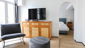 Ein modernes Hotelzimmer mit einem Flachbildfernseher, einem grauen Sofa, einem Holzkabinett und einem Bett mit blauer Kopfteil.