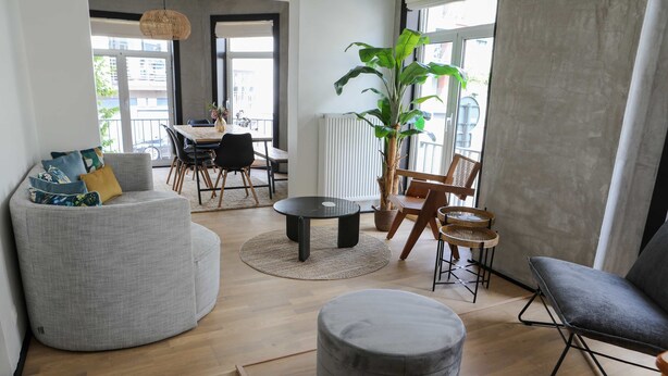 Ein modernes Wohnzimmer mit grauem Sofa, Hocker und einem Essbereich mit Tisch und Stühlen.