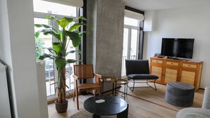 Ein modernes Wohnzimmer mit einer großen Pflanze, Holzstühlen, einem schwarzen Couchtisch, einem grauen Hocker und einem Fernseher auf einer Holzkommode.
