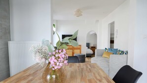Ein modernes Wohnzimmer mit einem Holzesstisch, einem Blumenstrauß, einem grauen Sofa und einem Fernseher im Hintergrund.