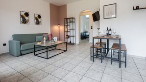 A modern living room with a sofa, coffee table, and dining table with chairs. There are framed pictures on the wall, a TV mounted above the table, and a bookshelf. - Philomena Apartment by Ionian Luxury (Kerkira)