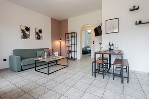 A modern living room with a sofa, coffee table, and dining table with chairs. There are framed pictures on the wall, a TV mounted above the table, and a bookshelf. - Philomena Apartment by Ionian Luxury (Kerkira)