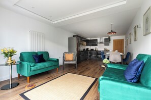 A modern living room with two green sofas, a small side table, and a dining area in the background.