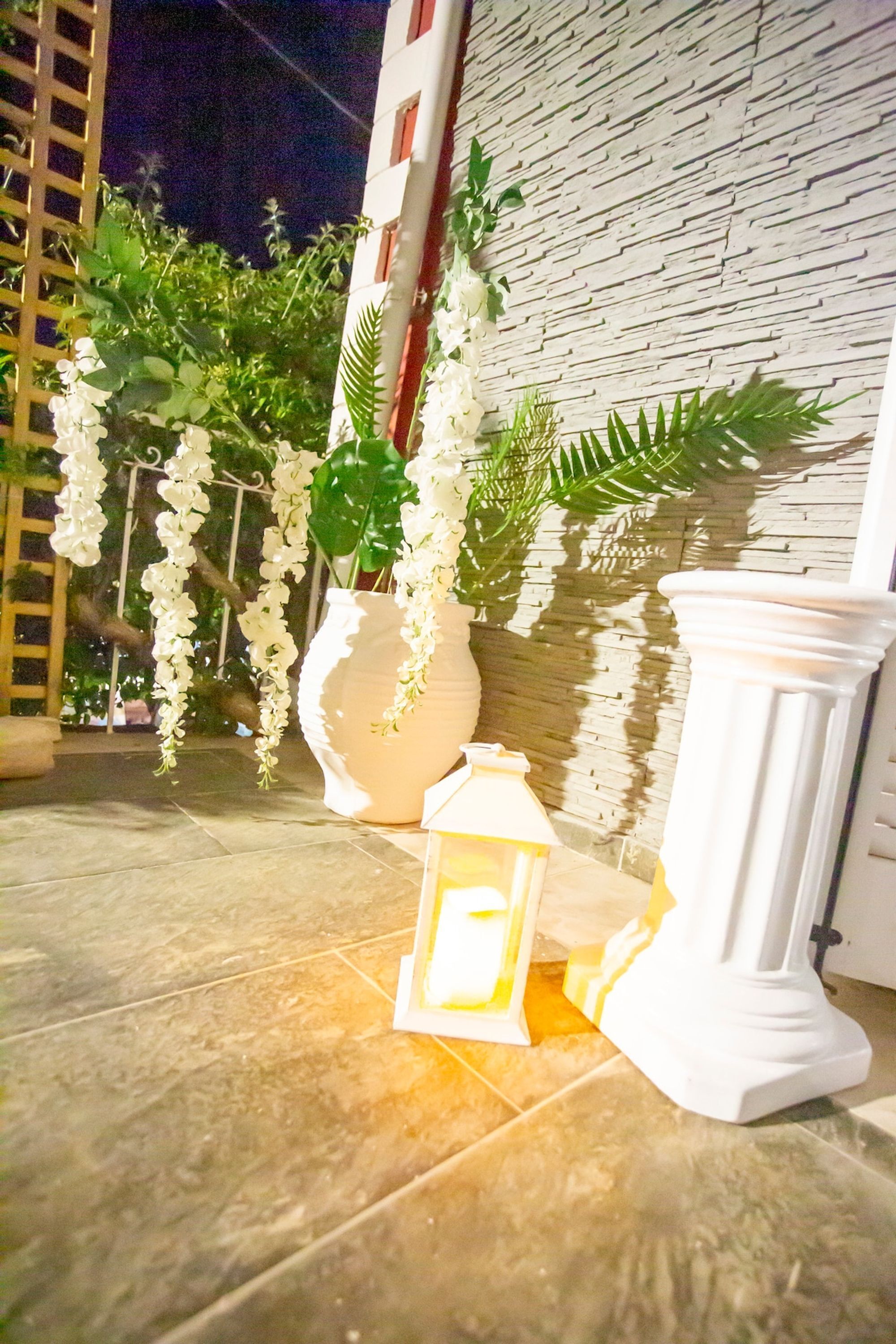 Un patio piastrellato con fiori bianchi pendenti, un lanterna e un pilastro bianco.