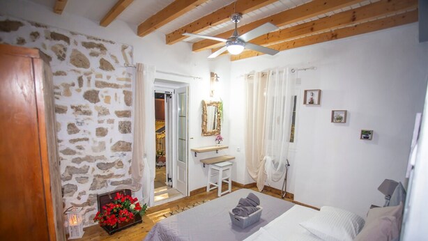 A bedroom with a bed, a wooden ceiling, a stone wall, a bathroom with a sink, and a mirror. - Archontissa House - Sea View (Mesongi)