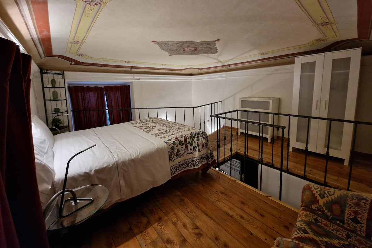A bedroom with a bed, a chair, a small table, a window with curtains, a ceiling with decorative elements, and a white wardrobe.