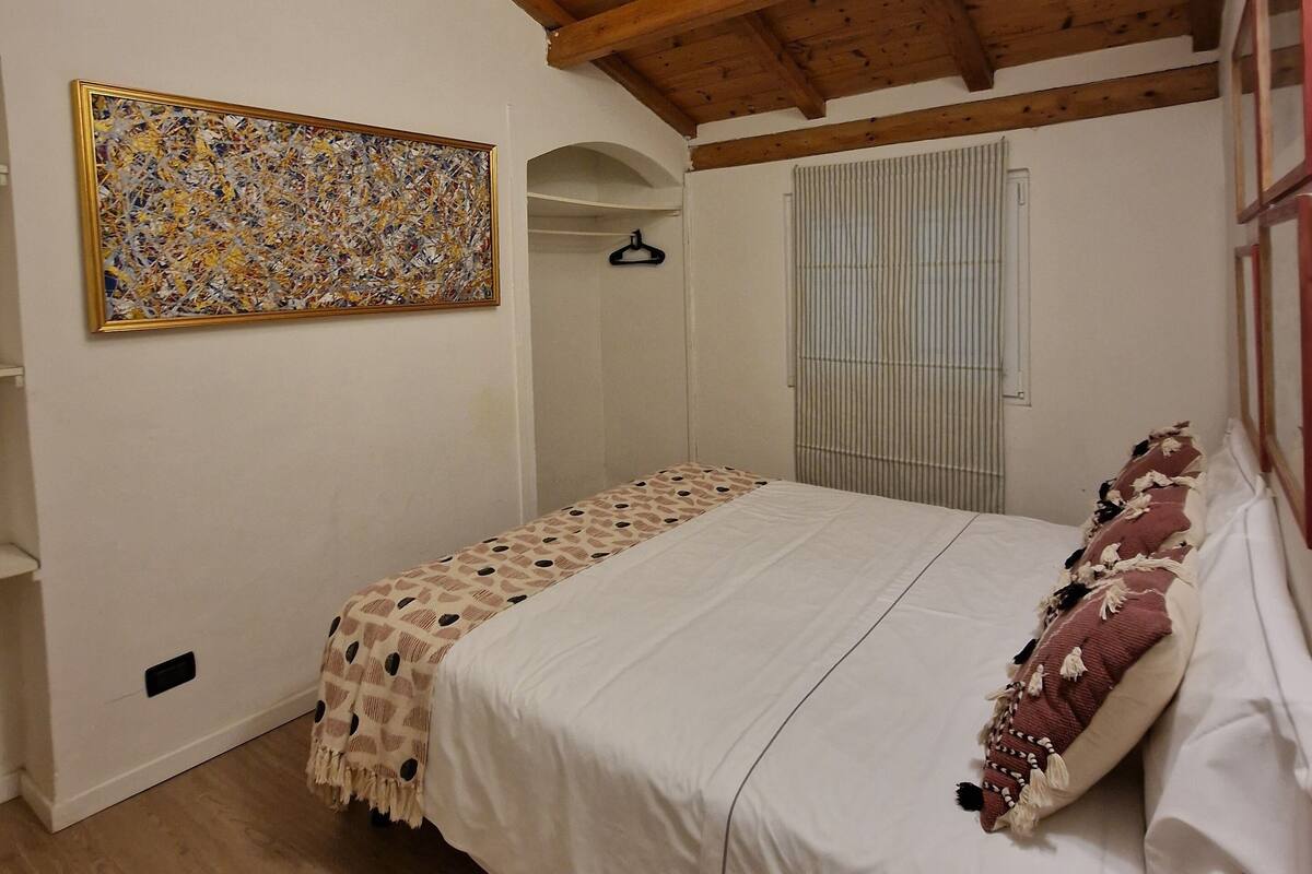 A bedroom with a wooden ceiling, a bed with a patterned bedspread, a hanging coat rack, and a window with blinds.