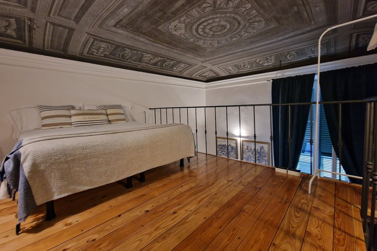 A bedroom with a wooden floor, a bed with a striped pillow, and a ceiling with intricate designs.