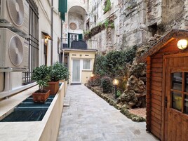 A narrow alleyway with a stone pathway, a wooden door on the right, and a building with a balcony and a door in the background.