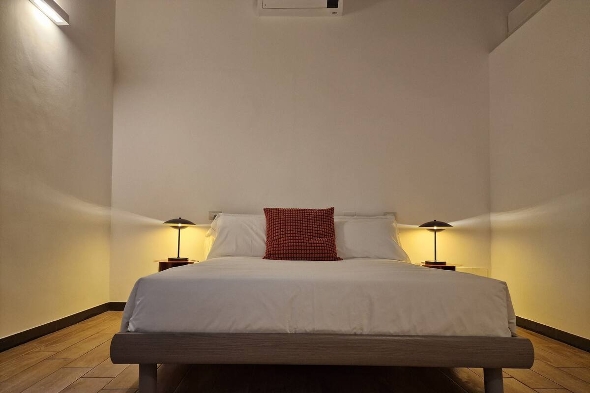 A neatly made bed with white bedding and a red patterned pillow, flanked by two bedside tables with lamps, in a room with wooden flooring and plain walls.