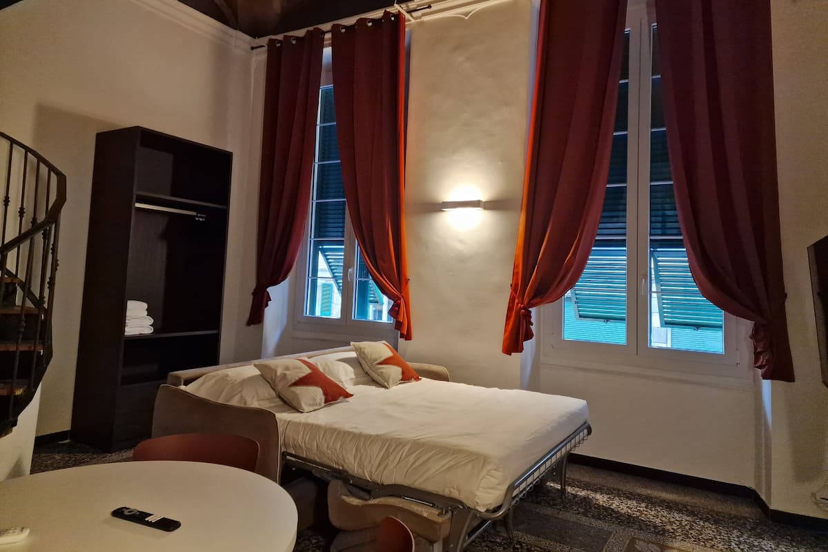 A hotel room with a bed, a desk, a chair, a bookshelf, and a window with curtains.