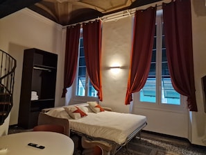 A hotel room with a bed, a desk, a chair, a bookshelf, and a window with curtains.